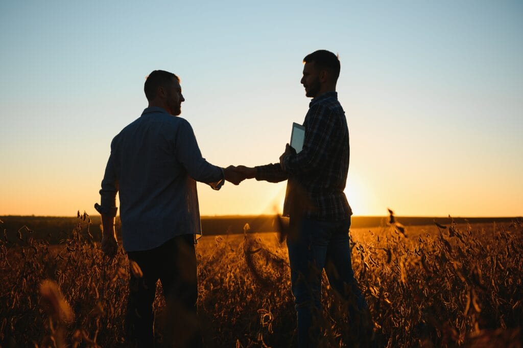Two farmers shaking hands in a soy crop field at sunset